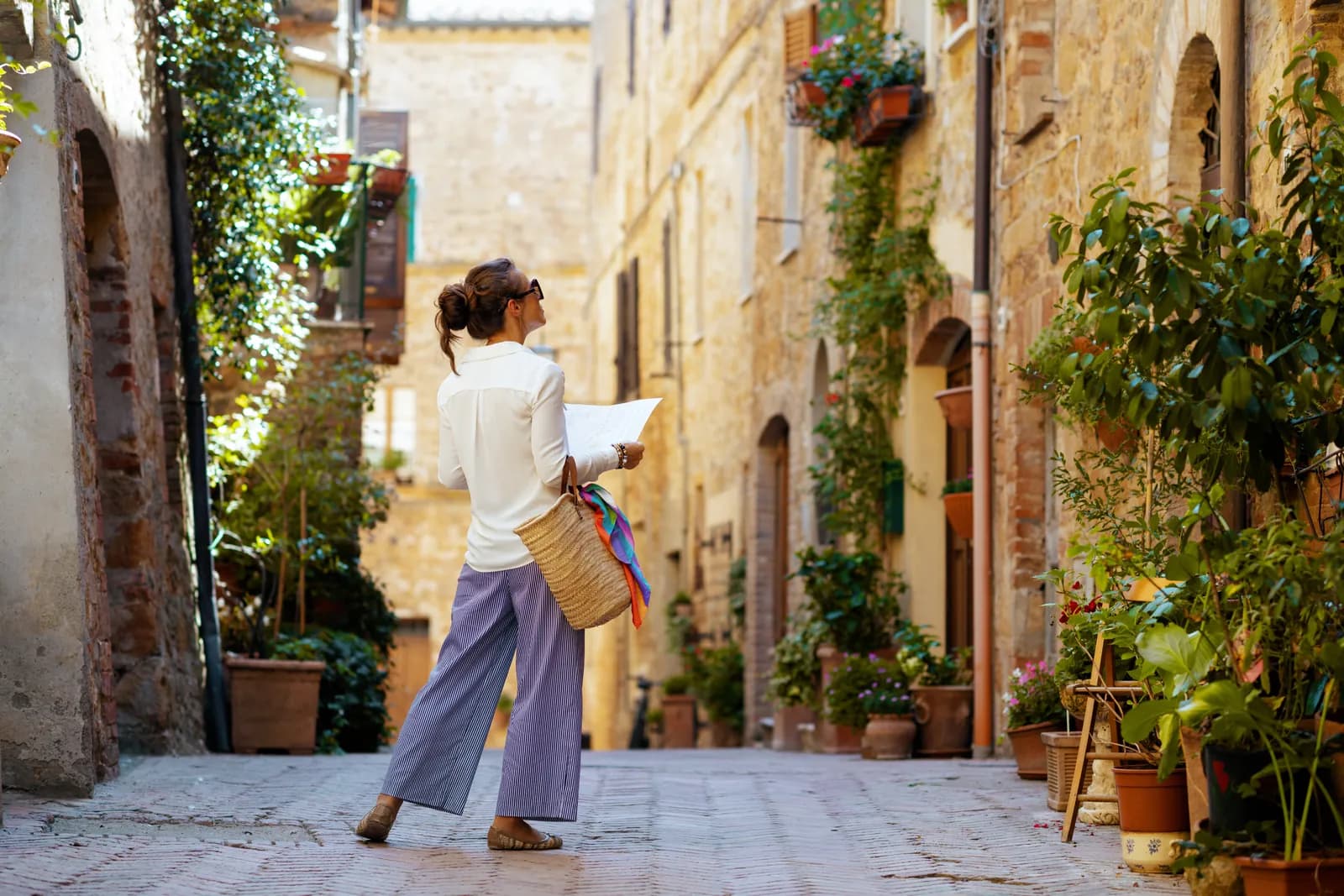 A student pauses in a sunlit Tuscan street, looking up at the stone buildings and the plants spilling from their balconies.